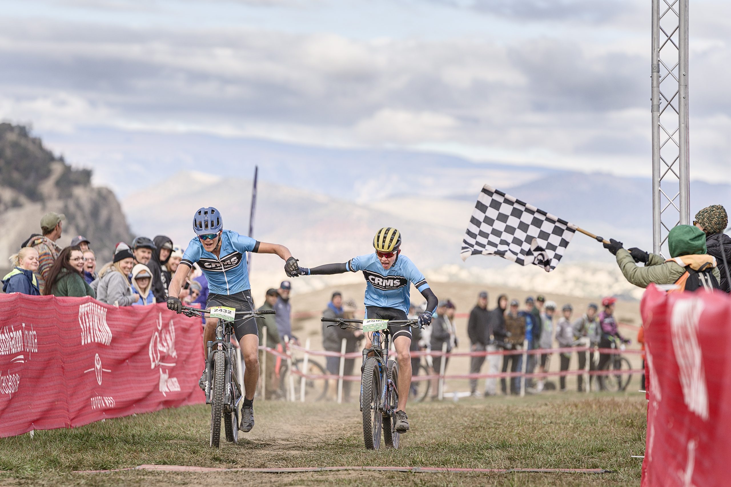 2 CRMS students holding hands as they ride their bikes across the finish line of a race.